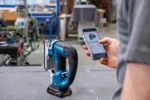 作業場で、スマートフォンを使ってコードレスジグソーを調整している人。.