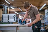 作業場では安全装備を身に着けた人がジグソーを使って木の板を切っています。