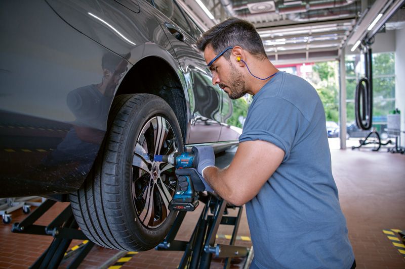 安全装備を身に着けた人がインパクトレンチを使って車のホイールのボルトを締めています。.