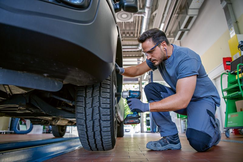 安全装備を身に着けた人が、コードレスインパクトレンチを使用して自動車のホイールボルトを締めています。.