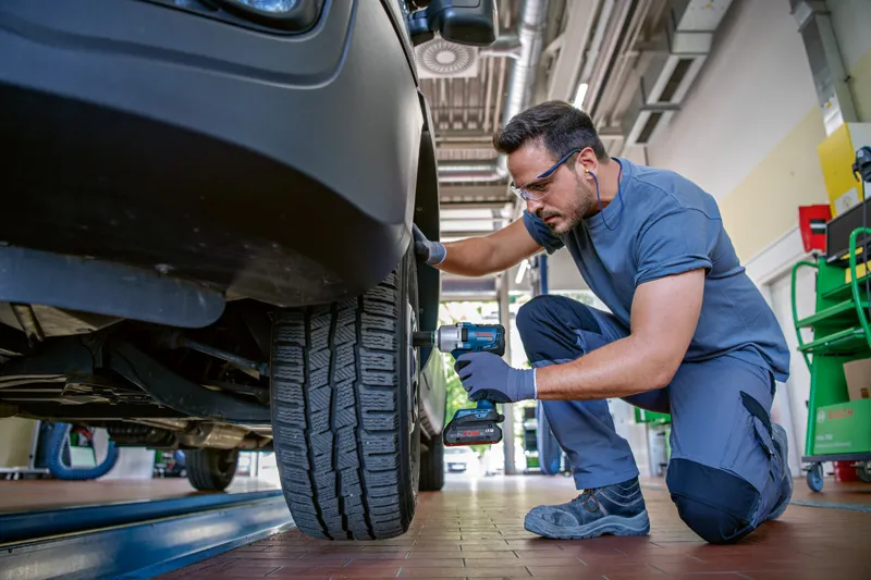 安全装備を身に着けた人が、コードレスインパクトレンチを使用して自動車のホイールボルトを締めています。.