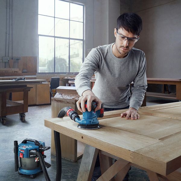 安全装備を身に着けた人が、接続されたオービタルサンダーで木製のドアを研磨しています。.