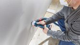 安全装置を装着した人が、コードレス乾式壁ルーターを使用して乾式壁に穴を開けています。.