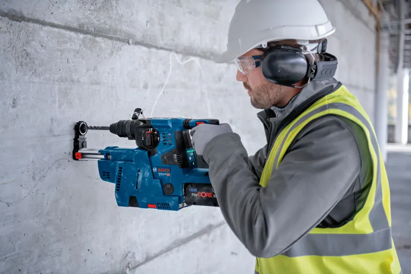 安全装置を装着した人が回転ハンマーを使用してコンクリートの壁に穴を開けています。.