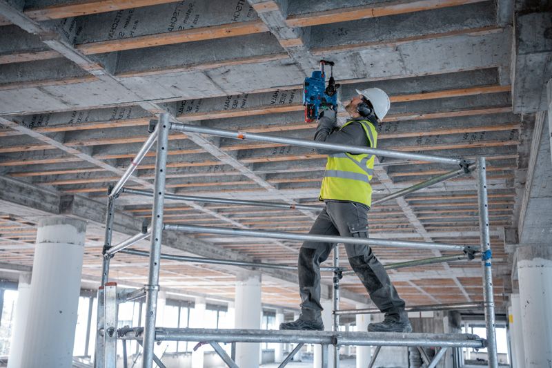 安全装備を身に着けた人が足場の上に立ち、コンクリートの天井でコアドリルを使用しています。.