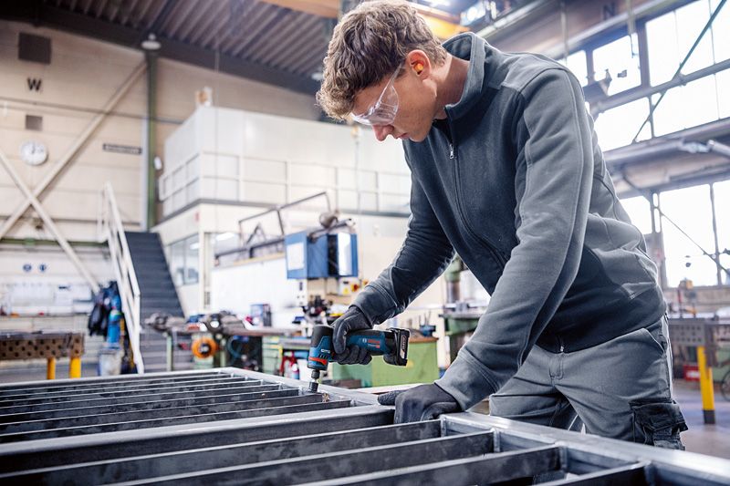 安全装置を装着した人が作業場で電動工具を使って金属の梁を固定しています。.