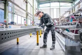 安全装備を身に着けた人が作業場で大きな金属の梁に穴を開けています。