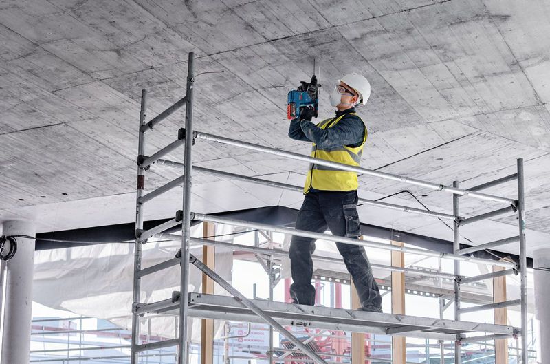 安全装備を身に着けた人が足場の上に立ってコンクリートの天井に穴を開けています。.