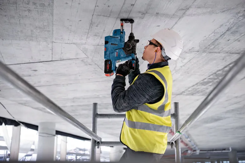安全装置を装着した人が電動工具を使用してコンクリートの天井に穴を開けています。.