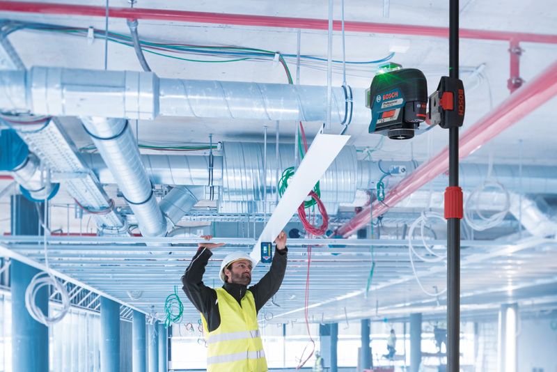 安全装備を身に着けた人が、伸縮ポールにレーザーレベルツールを使って天井パネルを設置しています。