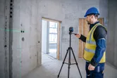 安全装置を装着した人がコンクリートの部屋で三脚の上のレーザーレベル測定ツールを操作しています。
