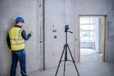 安全装備を身に着けた人が三脚に取り付けたレーザーレベルツールを使ってコンクリートの壁に印を付けています。