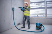 安全装備を身に着けた人が、コードレス集塵機を取り付けた状態でコンクリートに穴を開けています。.