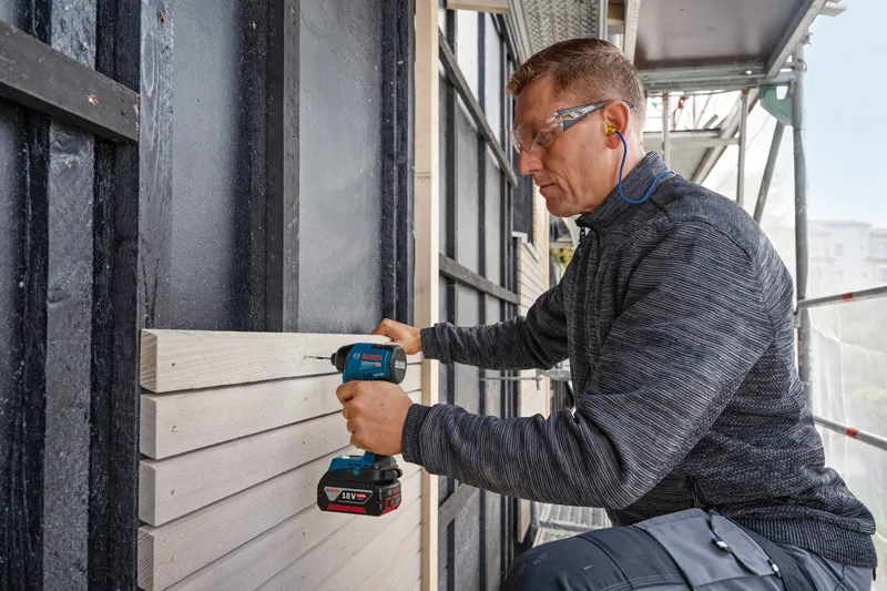 安全装備を身に着けた人がコードレスドリルで木製パネルを壁に固定しています。.