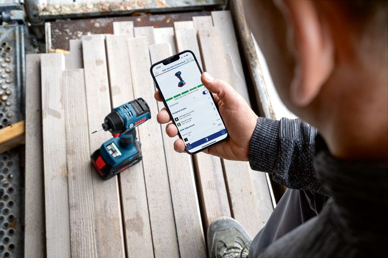 コードレスドリルの近くで、スマートフォンで電動工具のアプリ機能をチェックしている人。.