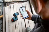 コードレスドリルの近くで、スマートフォンで電動工具のアプリ機能をチェックしている人。