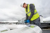 安全装備を身に着けた人が屋外の大きな金属ダクト上で電動ドリルを使用しています。.