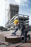 安全装備を身に着けた人が、建設現場でコードレスのこぎりを使って合板を切っています。