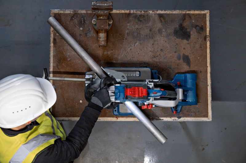 安全装置を装着した人が作業台の上で電動のこぎりを使って金属パイプを切断しています。.