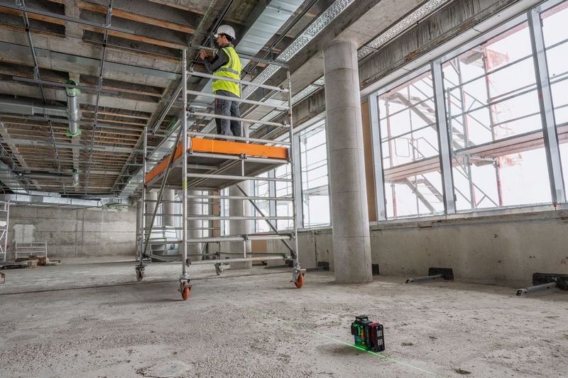 安全装備を身に着けた人が、レーザーレベルツールで線を投影しながら足場の上で作業しています。.