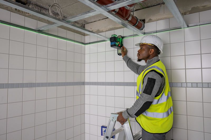 安全装備を身に着けた人がはしごからタイル張りの壁にレーザーレベル測定ツールを設置しています。.