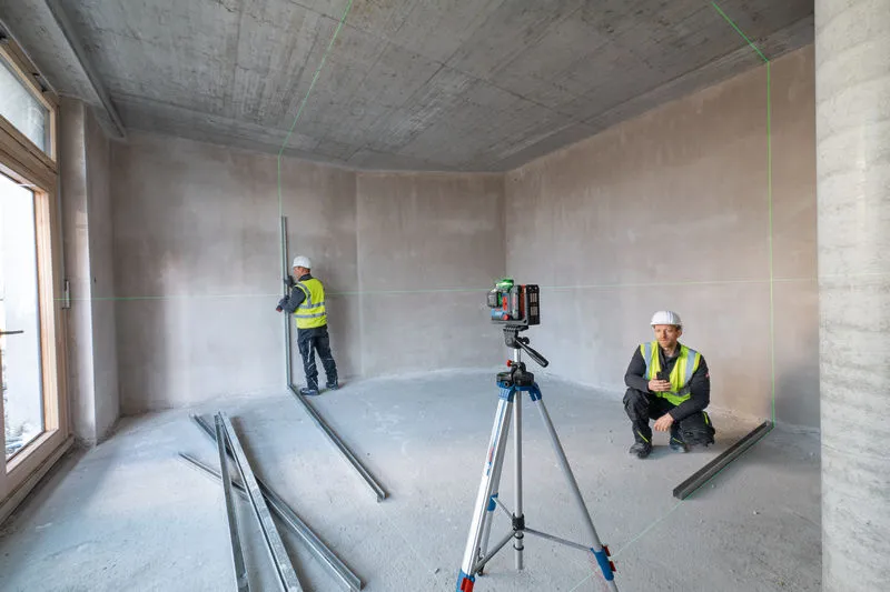 安全装備を身に着けた2人が未完成の部屋でレーザーレベルツールを使用しています。