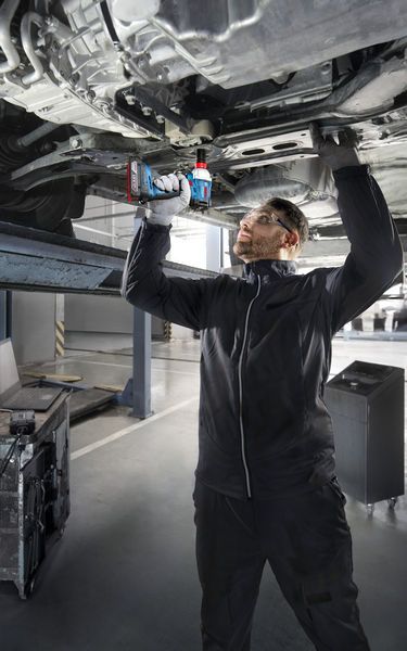 安全装置を装着した人がリフトされた車両の下でコードレスドリルを使用しています。.