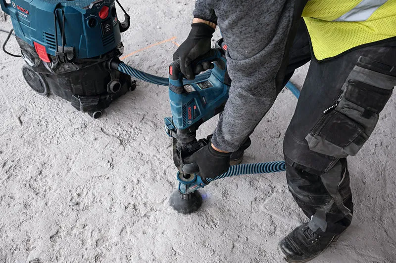 安全装備を着用した人が、SDS マックスの集塵装置を使用してコンクリートに穴を開けます。.