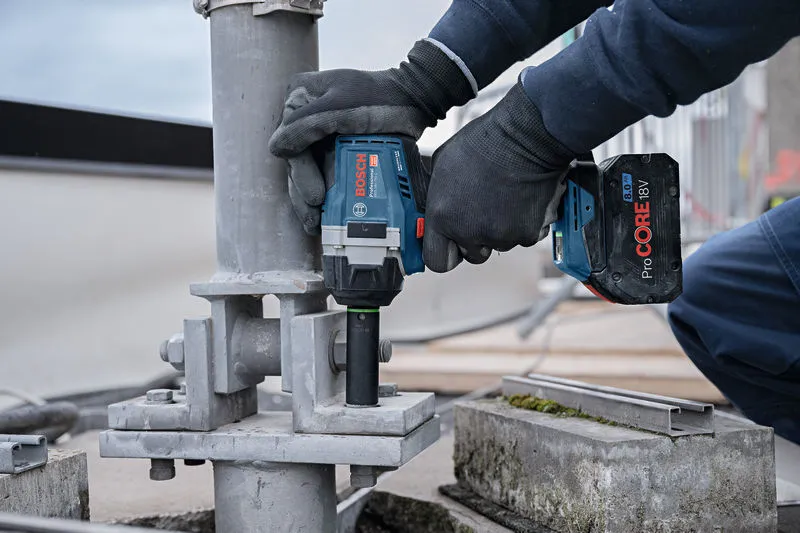 安全装備を身に着けた人が、コードレスインパクトレンチを使用して金属構造物に大きなボルトを締めます。.