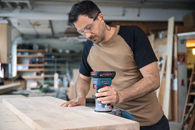 安全装備を身に着けた人が、コードレスパームルーターを使用して木の板の端をルーティングしています。.