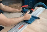 人が木製の作業台上のガイド レールに沿ってコードレス パーム ルーターを操作しています。