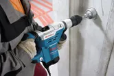 安全装備を身に着けた人が回転ハンマーでコンクリートの壁に穴を開けています。.