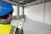 安全装備を身に着けた人が何もない部屋の中でスマートフォンを使ってレーザーレベルツールを調整しています。.