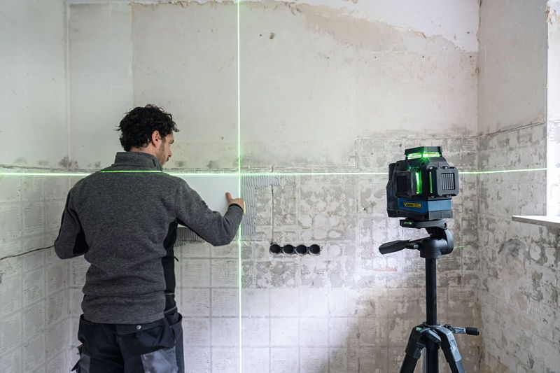 三脚に取り付けた緑色のレーザーレベル測定ツールを使用して、壁のタイルの位置を合わせている人。.