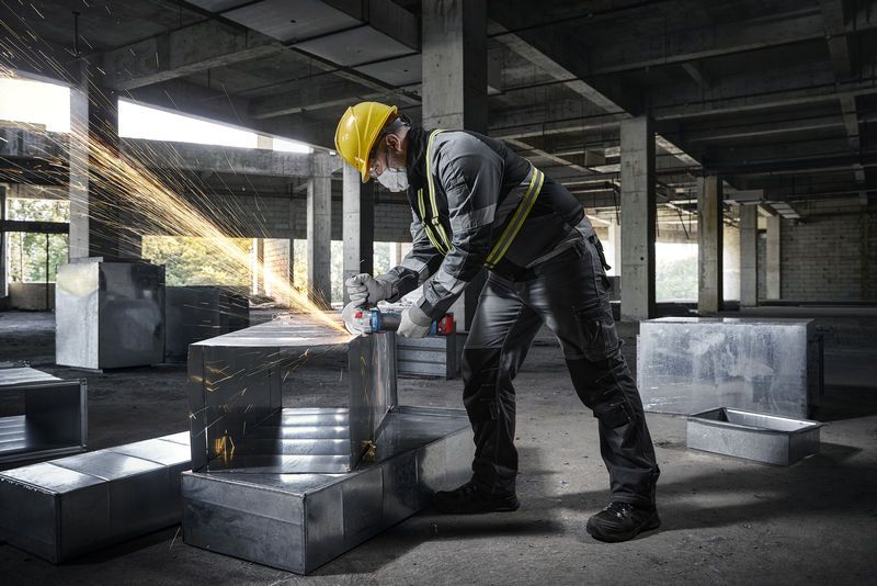 建設現場で、安全装備を身に着けた人が金属ダクトの上でアングルグラインダーを使用しています。.