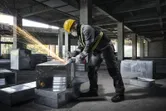 建設現場で、安全装備を身に着けた人が金属ダクトの上でアングルグラインダーを使用しています。.