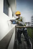 安全装備を身に着けた人が足場から建物の壁に穴を開けています。