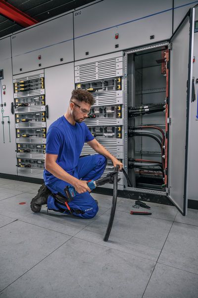 GKH 18V-50 安全装備を身に着けた人が、開いた電気パネルの前で太いケーブルを切断しています。.