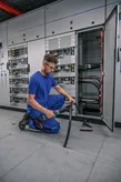 安全装備を身に着けた人が、開いた電気パネルの前で太いケーブルを切断しています。