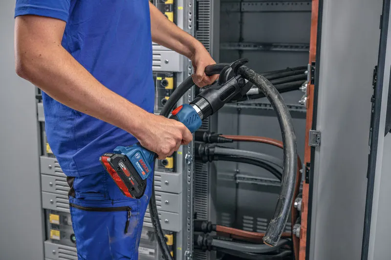 安全装置を装着した人が、コードレス油圧切断工具を使用して太いケーブルを切断しています。.