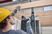 安全装備を身に着けた人が木の梁にネジをドリルで打ち込んでいます。
