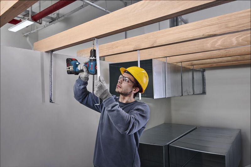 安全装備を身に着けた人が木の梁の下の金属棒にドリルで穴を開けています。.