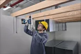 安全装備を身に着けた人が木の梁の下の金属棒にドリルで穴を開けています。
