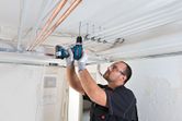 安全装置を装着した人が天井に穴を開けて電気配管を設置しています。.