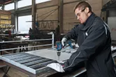 安全装備を身に着けた人が工場の作業台で金属メッシュの穴あけ作業をしている。.
