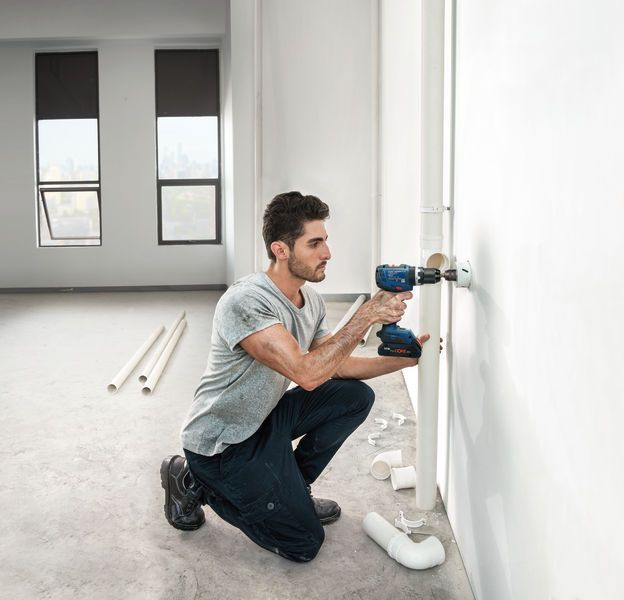 明るい未完成の部屋で、配管を設置するために壁に穴を開けている人。.