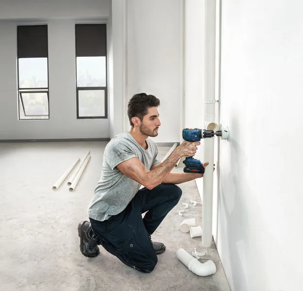 明るい未完成の部屋で、配管を設置するために壁に穴を開けている人。.