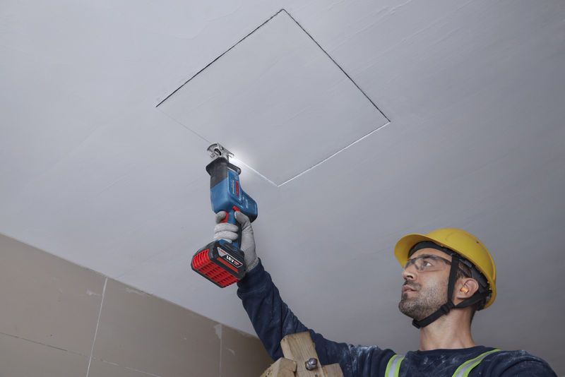 安全装置を装着した人がレシプロソーで天井パネルを切断しています。.