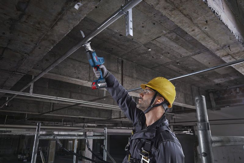 安全装置を装着した人が往復鋸を使用して頭上の金属パイプを切断しています。.