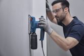 安全装備を身に着けた人が白い壁の上でオービタルサンダーを使用しています。.